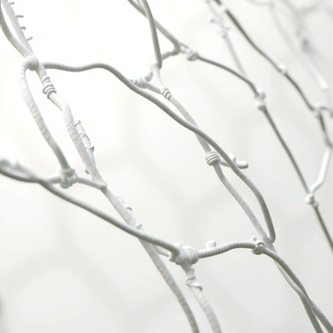 A close-up image of a soccer goal net, showcasing its intricate texture. The net is made of white nylon ropes, intricately woven together in a diamond pattern. The fibers appear taut and slightly shiny, reflecting light in various areas. The background is blurred, focusing attention on the fine details of the net. Small strands are visible, with some fraying edges, giving a sense of wear from use. The photo conveys a feeling of being up close to the action, emphasizing the net's role in the game.