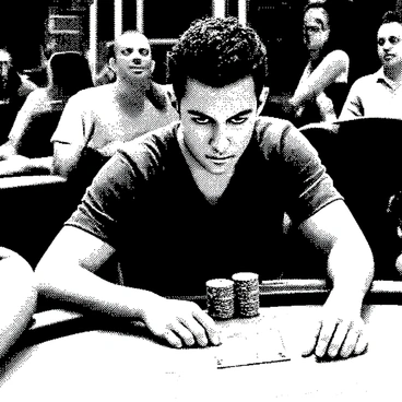 A blackjack player is seated at a casino table, intently focused on the cards in front of him. He has short dark hair and a contemplative expression on his face, conveying deep concentration. His hands are resting on the table, partially covering a stack of chips. The surrounding environment includes other players engaged in the game and a dealer in the background, all contributing to the lively atmosphere of the casino. The lighting is bright, emphasizing the tension of the moment.