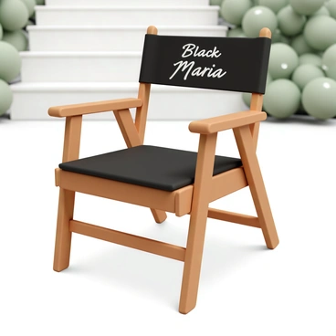 A folding director's chair with a wooden frame and black fabric. The chair features a backrest that is elegantly embroidered with the text "Black Maria" in bold white letters. The seat is wide and comfortable, and the arms of the chair are smooth and polished. The chair is positioned on a simple outdoor patio with a soft focus on greenery in the background, showcasing the chair's stylish design and craftsmanship.