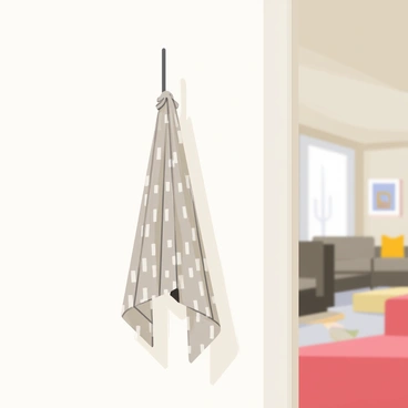 A baluchon, which is a simple fabric bundle, hangs from a sleek, metallic wall hook. The baluchon is made of grey and white patterned fabric, slightly wrinkled, suggesting it is filled with belongings. The wall behind the hook is a smooth, light-colored surface, emphasizing the simplicity of the space. The modern apartment features a clean, minimalist aesthetic with sparse decor, and in the background, a soft glimpse of a contemporary living room can be seen with muted colors.