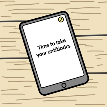 A smartphone with a sleek design is positioned on a wooden table. The screen displays a bright notification that reads, "Time to take your antibiotics." The app icon is visible in the corner, and the background shows a subtle texture of the wooden surface. The smartphone is slightly angled to provide a clear view of the notification without any distractions.