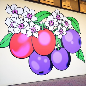 A large mural on the wall of a downtown café features vibrant plums in shades of purple and red. Surrounding the plums are delicate white tree blossoms with soft pink centers. The composition includes green leaves interspersed among the fruits and flowers. The café wall serves as the backdrop for this colorful display, enhancing the atmosphere of the space.