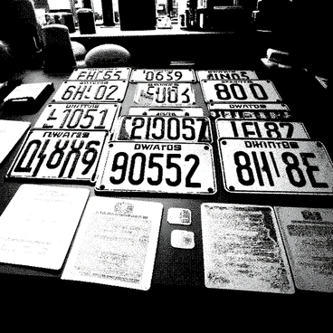 An assortment of old registration plates is neatly arranged on a wooden desk inside a shop. The registration plates display various numbers and letters, showcasing their vintage designs. Scattered around the plates are a few weathered documents, resembling old vehicle registrations, with faded text and stamps. The desk is slightly cluttered, adding to the atmosphere of a shop filled with history. Natural light filters in from a nearby window, casting soft shadows on the surfaces.