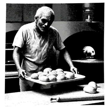A zombie baker with decaying skin and tattered clothing is standing next to a large industrial oven. He has disheveled hair and hollow eyes that reveal a lack of life. In his bony hands, he is pulling a tray filled with freshly baked bread, the golden crusts glistening under the light. The scene includes flour scattered across a wooden countertop, with some bread rolls cooling nearby. A rolling pin and a few baking tools are visible, adding to the atmosphere of a busy bakery.