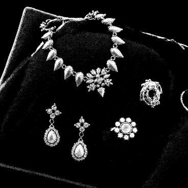 A set of intricate jewelry pieces is arranged on a cushion of black velvet. The jewelry includes a necklace with detailed metalwork, a pair of ornate earrings, and a ring, each featuring precious gemstones. The gemstones are emitting a soft, ominous glow, illuminating the surrounding area. The velvet display is smooth and rich in texture, contrasting with the sharp, angular designs of the jewelry pieces. Shadows play across the velvet, enhancing the eerie atmosphere surrounding the cursed items.