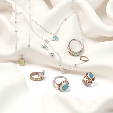 A collection of handcrafted jewelry, featuring several intricate necklaces and rings. The necklaces have delicate chains with various pendants, including gemstones and metal designs. The rings are adorned with colorful stones, intricate filigree work, and unique shapes. The jewelry is artistically arranged on a soft, textured fabric that creates an elegant backdrop, enhancing the beauty and details of each piece.
