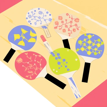 A wooden table with a smooth surface, showcasing a collection of uniquely designed table tennis paddles. Each paddle features a different vibrant color scheme and intricate patterns. Some paddles have geometric designs in shades of blue and yellow, while others display floral motifs in red and green. The handles of the paddles vary in style, with some being wrapped in black grips and others having a natural wood finish. The background includes a subtle hint of a sports venue, creating an atmosphere of competition.