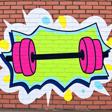 A large barbell is illustrated in bold colors on a brick wall. The barbell has bright red weights at each end and a shiny black central bar. Surrounding the barbell are dynamic splashes of color including green, blue, and yellow, creating a lively background. The graffiti mural includes abstract shapes and patterns that enhance the energetic feel of the scene. The wall has a rough texture, showcasing the urban environment.