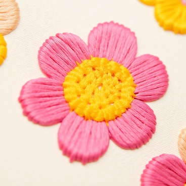 A close-up view of an embroidered flower on fabric. The flower features vibrant petals in shades of pink and yellow, with intricate stitches that showcase the craftsmanship. The fabric underneath is a soft cream color, allowing the bright colors of the embroidery to stand out clearly. Fine details in the stitching can be seen, with varying thread thicknesses adding depth and texture to the design. The background is softly blurred to emphasize the flower's vivid details.